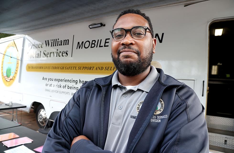 Damien Quesada stands in front of the Prince William Social Services mobile unit, wearing county-branded attire with arms crossed.