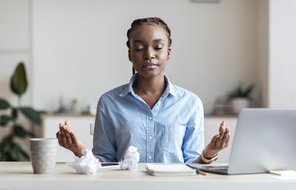 A woman sitting at a desk in front of a laptop, with her eyes closed and hands raised in a calming, meditative pose.