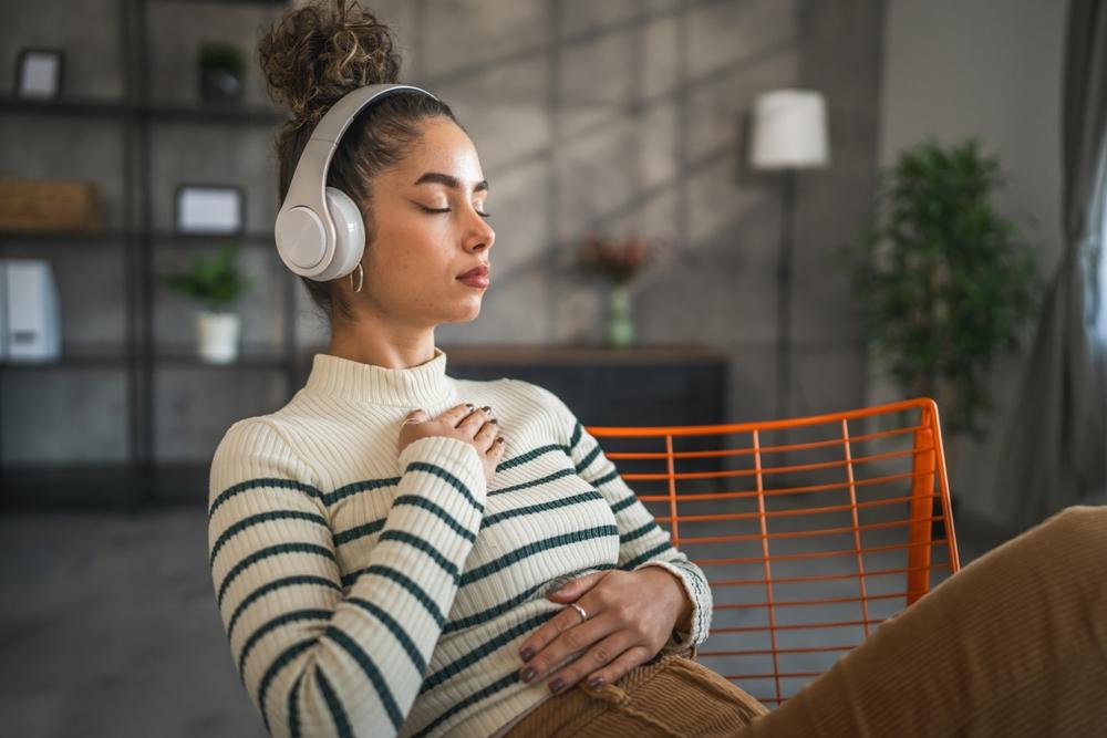 A woman sitting on a chair, wearing white headphoneswith her eyes clothes and hands resting gently on her chest and stomach in a calming, mindful pose. 