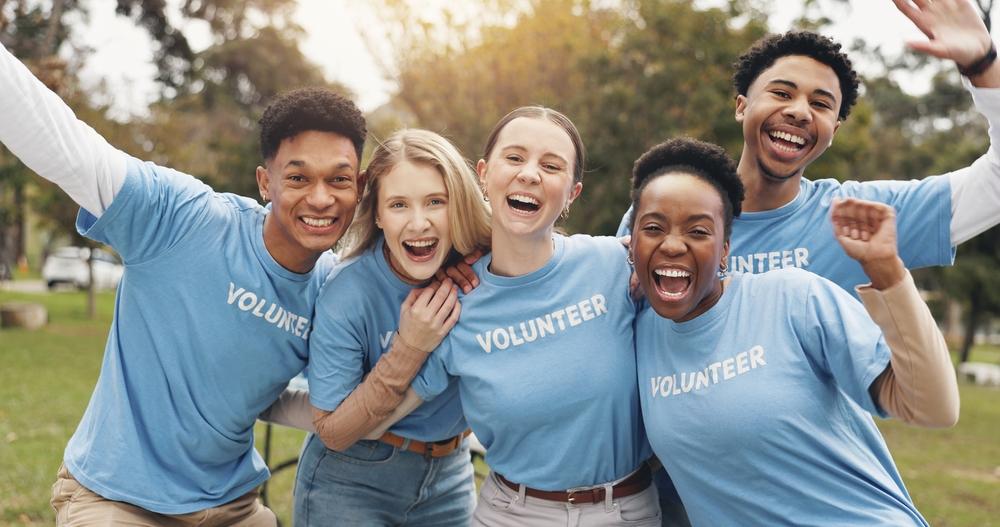 A diverse group of four young adults wearing matching blue t-shirt, standing outdoors and smiling warmly with their arms around each other. 