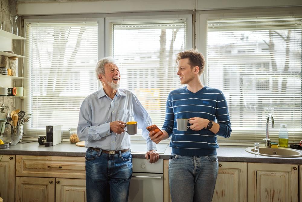 Two men, one older and one younger, standing together in a brightly lit kitchen holding coffee mugs and engaging in a supportive conversation. 